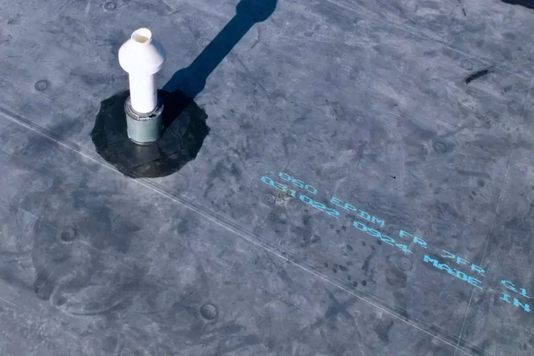 White vent pipe sealed into a dark gray EPDM flat roof membrane with printed manufacturer markings.