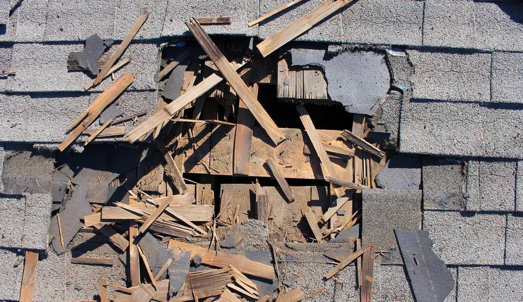 Hole torn through a shingle roof with broken decking boards and shingle debris around it
